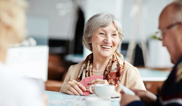 Smiling older woman enjoying playing cards around a table with others with a cup of tea